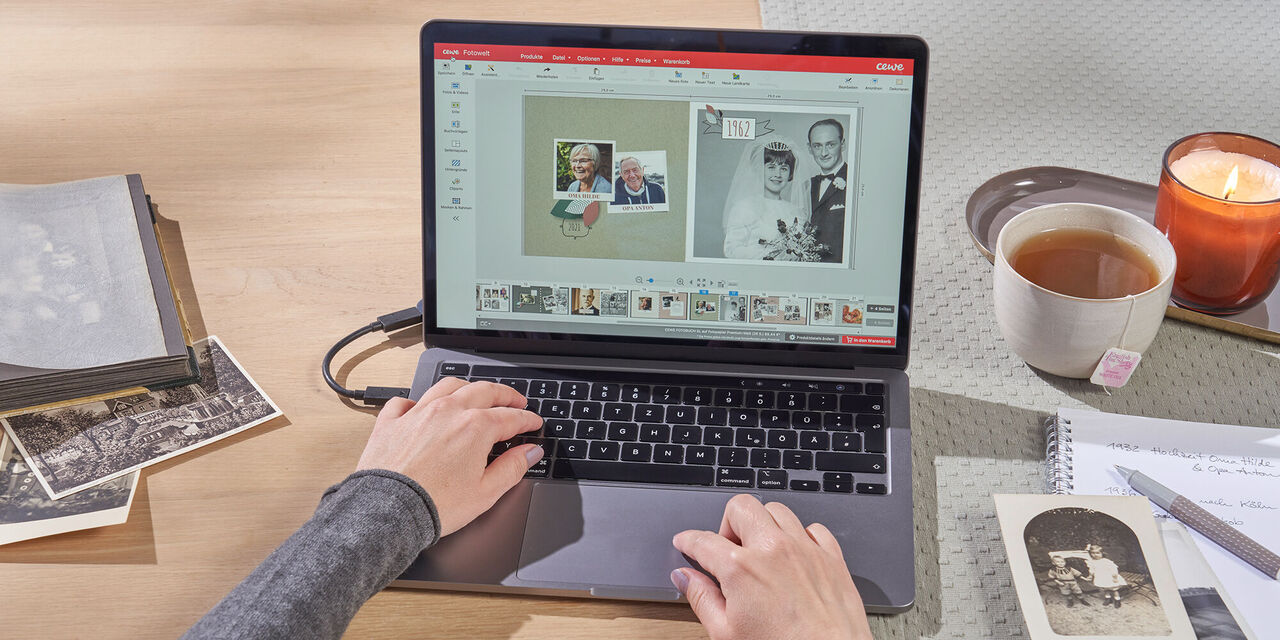 Ein Laptop steht auf einem Tisch. Auf dem Display ist die CEWE Fotowelt Software geöffnet, in der eine Person ein Fotobuch mit der Familienchronik-Buchvorlage bearbeitet. Links neben dem Laptop liegen analoge Schwarzweiß-Fotos. Rechts stehen eine Tasse Tee und eine Kerze.