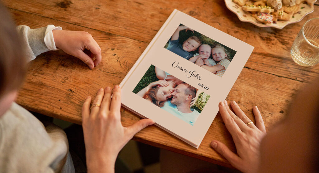 Blick über die Schulter zweier Betrachter: Auf einem Holztisch liegt ein geschlossenes CEWE FOTOBUCH. Auf dem Cover steht in goldener Schrift "Unser Jahr". Es sind zudem zwei Bilder der Familie zu sehen.