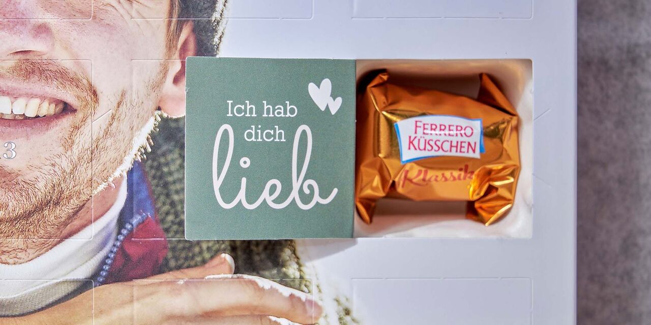 Auf der Innenseite einer geöffneten Tür des Adventskalenders steht “Ich hab dich lieb”. Daneben sind zwei kleine Herzen zu sehen. Hinter der Tür steckt ein Ferrero Küsschen.