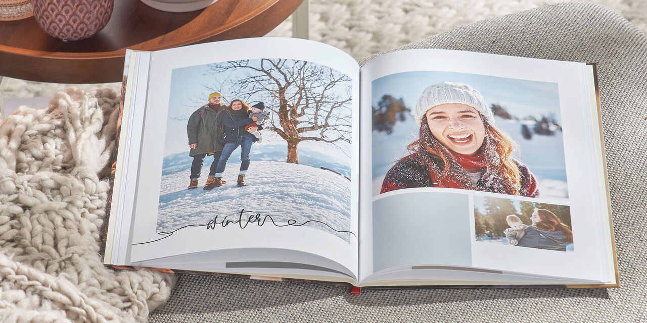 Auf einem grauen Sofa liegt ein aufgeschlagenes CEWE FOTOBUCH. Darin sind mehrere Fotos einer Familie sowie das Wort „Winter“ zu sehen. Links daneben steht ein Tisch mit Teelichtern und einer Pflanze.